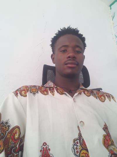 A young adult male is depicted seated, looking directly forward, in the city of Agingi Zion, Nigeria. He has dark skin, short dark curly hair, and a mustache. He wears a white long-sleeve button-down shirt featuring a multi-colored paisley pattern in shades of red, orange, yellow, and brown. A dark beaded necklace with a small dark pendant is visible around his neck. He is positioned in a black office chair with a mesh backrest. The background consists of a plain white wall, which exhibits minor cracks and discoloration.
