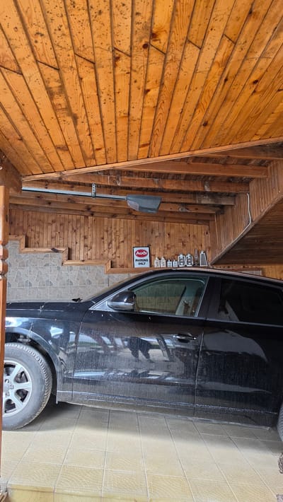 A black car is parked in a garage with a wooden-paneled ceiling and walls. The garage floor is tiled. A sign that reads "AUDI PARKING ONLY" is affixed to the wall. The car appears dusty. The scene is consistent with a residential garage in Rogachevo, Bulgaria. No people are visible. The car is the primary subject. The presence of Audi-branded signage could be relevant if the garage is part of a business, car dealership, or a private residence with an affiliation to the Audi brand.