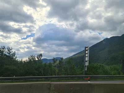 The image depicts a view from a vehicle on a highway in Schottwien, Austria. The scene is dominated by a dramatic cloudy sky with patches of blue. A dense, dark green forest covers the slopes of a large mountain to the right of the frame. A road barrier runs horizontally across the foreground, separating the highway from a grassy verge. A tall signpost with multiple reflective panels is visible on the right side of the road. The overall atmosphere suggests a natural, mountainous landscape.