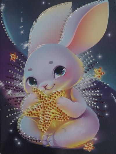 This image depicts a close-up, high-angle view of a diamond painting of a cartoon bunny holding a star. The bunny is pastel purple and yellow, with large blue eyes and pink cheeks. It is seated and holding a large, yellow, diamond-encrusted star in its paws. The background is a dark blue night sky with scattered stars and streaks of light. The edges of the bunny's ears and outline are decorated with clear, red, and dark red diamonds. There are also several small, diamond-encrusted stars scattered around the bunny. The image does not contain any information about Nizhneye Myachkovo, Russia.