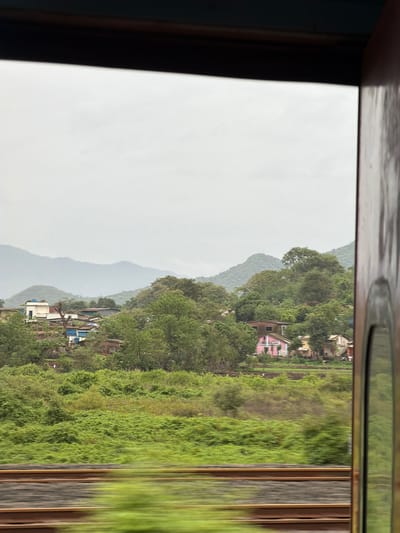 The image captures a view from a moving train window, showing a village nestled amidst lush green hills under a cloudy sky. In the foreground, blurred train tracks run horizontally across the frame. Beyond the tracks, dense, vibrant green foliage covers the landscape, with a small village scattered on the slopes of the hills. The village consists of several houses, some with tiled roofs and brightly colored facades, including a distinctive pink house. In the background, layers of hazy, green mountains recede into the distance. The overall scene suggests a rural or semi-rural setting, likely in the outskirts of Vasai-Virar, India, characterized by its natural beauty and modest human settlements. The overcast sky indicates a potentially damp or humid climate.