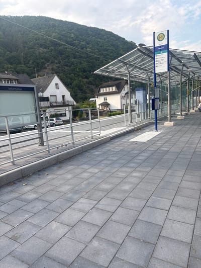 The image shows a bus stop in Ahrbrück, Germany. The bus stop is covered by a modern, transparent roof supported by metal beams. A blue sign with a green bus symbol indicates it is a public transportation stop. The sign lists destinations such as "Ahrbrück", "Bahnhof", and various other towns and stations, including "Remagen", "Nassau", and "Kirchahr". To the left of the bus stop, there is an information panel with the word "Information" visible. The ground in front of the bus stop is paved with grey rectangular tiles. In the background, there are houses with tiled roofs and a steep, tree-covered hillside. A white van and a silver car are parked on the street behind the bus stop. The sky is cloudy, and the sun is casting shadows on the pavement, suggesting it is daytime.