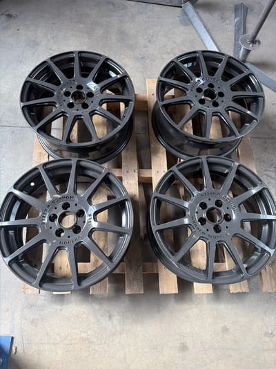 Four dark gray, multi-spoke alloy wheels are arranged on a wooden pallet on a concrete floor. The wheels appear to be new, with a metallic finish. Each wheel has "MONACO WHEELS" embossed around the center hub. The background consists of a concrete floor and some industrial metal structures. The setting is likely a workshop or storage area.