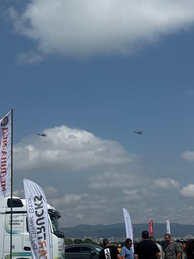 The image captures an outdoor event under a bright, partly cloudy sky, with a backdrop of distant mountains and urban buildings. The perspective is slightly elevated, looking towards the event area.
In the sky, two helicopters are visible; one flies higher against the clear blue, while the other is lower, partially obscured by a large white cloud.
At ground level, a group of people, predominantly men in casual attire, are gathered, seemingly observing the event. A prominent white truck, likely a semi-truck, is partially visible, adorned with promotional materials. Another dark vehicle is seen behind it.
Several promotional flags and banners are present. A large white banner in the foreground displays the mirrored text "TRUCKS" and "Silver Star." Another banner below it shows "DAM-AHIIDAM." Further back, smaller flags include one with "АТАР" and another with a red emblem. Text on the white truck includes "Mercedes-Benz Actros 600," "ОТВЕДЕН ЗА ПРОМЯНА" (Reserved for Change), and "SILVER STAR."
The scene suggests a public event, possibly a truck show, a promotional gathering, or an air display, given the combination of helicopters and commercial vehicles. It is daytime with good weather. The setting near mountains and a city places the event in Madzherito, Bulgaria.