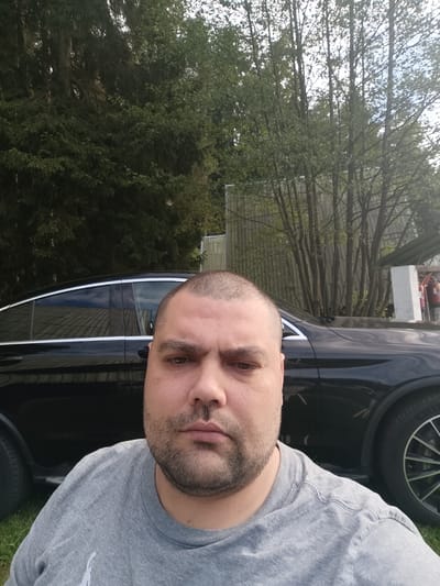 A close-up, head-and-shoulders selfie shows a man with short hair, a beard, and a grey t-shirt. He is looking directly at the camera with a neutral expression. Behind him, a black car is parked. The background consists of trees and a building. The scene appears to be outdoors, possibly in a suburban or rural area, and is not identifiable as being in the city of Babylon, Czechia.