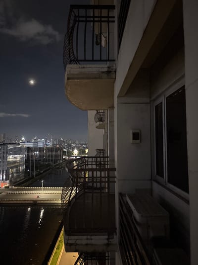 The image is a night-time view from a high-rise building in Pasay, Philippines. A sliver of the crescent moon is visible in the dark sky, with some clouds. Below, a cityscape unfolds with numerous buildings, some illuminated by lights, suggesting a densely populated urban area. A body of water, likely a river or bay, reflects the city lights. A road runs alongside the water, with a single vehicle visible on it, its headlights illuminating the path. The foreground shows a balcony railing, partially obscuring the view, and a portion of the building's exterior, including a window and an air conditioning unit. The overall impression is of a dynamic, nocturnal urban environment.