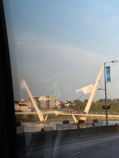 The image captures a reflection of the Peace Bridge in the city of Derry/Londonderry, United Kingdom, viewed through a window. The bridge is a prominent pedestrian and cyclist bridge spanning the River Foyle. The reflection shows the iconic white cable-stayed design of the bridge with its two inclined pylons.

In the background, there are modern buildings, suggesting an urban setting. The sky is partly cloudy, with a soft, diffused light. There are small, blue lights visible in a line across the sky, possibly from a decorative lighting feature or a reflection.

On the bridge itself, several people can be seen walking, indicating it is open and being used. Flower baskets hang from a lamppost on the right side of the image, adding a touch of color to the scene. The foreground shows a dark, reflective surface, consistent with a window, with some smudges or reflections visible. A road surface with lane markings is also visible in the lower part of the image, implying the viewpoint is from a vehicle or near a road. The overall impression is a peaceful, urban scene bathed in gentle light, with the Peace Bridge as the central architectural element.