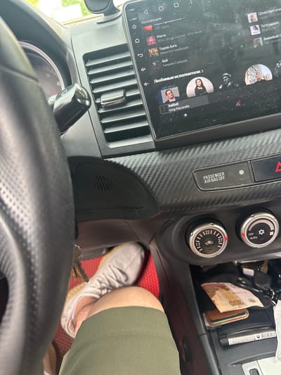 A first-person view from inside a car shows a person's left leg and foot, clad in a green shorts and a white sock, resting on the floor. The car's interior features a carbon fiber-patterned dashboard, a touchscreen infotainment system displaying a music app with Russian text and song titles such as "Mama" and "Guli", and climate control knobs. Keys are visible near the steering column. Outside, sunlight suggests a daytime setting, possibly in Kuzmolovskiy, Russia, given the presence of Russian currency, a 100 ruble banknote, in the center console. The car appears to be parked.