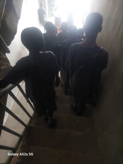 A group of children, silhouetted against a bright light source at the top of a staircase, are descending. The staircase has concrete steps and a metal railing on the left side. The environment appears to be an indoor or semi-enclosed space with rough, textured walls. The children are wearing dark clothing. The image is tagged with "Galaxy A52s 5G".