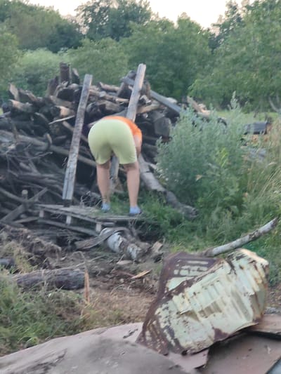 The image depicts a person, seen from behind, wearing light green shorts and an orange shirt, bending over near a pile of wood. The person is wearing blue shoes. The background consists of trees and dense green foliage. In the foreground, there is a large piece of rusted metal. The scene appears to be outdoors, possibly in a rural or overgrown area. The location is stated as Kochmar, Bulgaria.