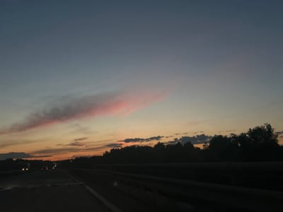 The image depicts a view from a vehicle traveling on a highway near Starosemeikino, Russia at dusk or dawn. The sky is a gradient of colors, transitioning from a darker blue at the top to shades of orange and pink near the horizon. A pink cloud is visible in the upper-left. A line of trees silhouettes the horizon. On the left side of the road, there are faint headlights suggesting other vehicles in motion. The highway itself is dark and reflective, and the guardrail is visible on the right.