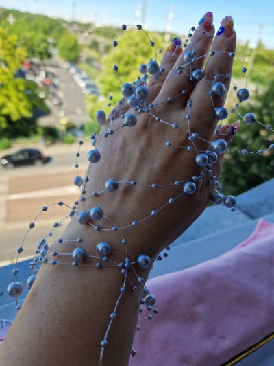 A person's hand, adorned with a beaded strand of gray pearls, rests on a pink fabric. The hand is positioned in front of a blurred outdoor scene with trees, a street with cars, and distant structures under a clear sky. The person's fingernails are decorated with colorful floral nail art. The setting appears to be a balcony or an elevated vantage point. The image was taken in the city of Fresnes, France.