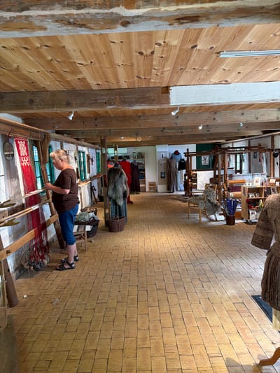 The image depicts the interior of the Sagnlandet Lejre museum in Lejre, Denmark, specifically the textile workshop area. A middle-aged woman, wearing a brown t-shirt and blue jeans, is actively engaged in weaving on a large wooden loom, her hands manipulating the threads. The loom is set up with vibrant red and white threads, indicating ongoing textile work.
The workshop environment is rustic, with exposed wooden beams and a natural wood plank floor. Various other looms and textile-related equipment are visible throughout the space, along with baskets of raw materials, possibly wool. Displayed on mannequins and shelves are finished textile products and garments, showcasing traditional craftsmanship. The background reveals more of the museum's interior, with windows and doorways leading to other sections. The lighting appears to be a mix of natural light from the windows and artificial lighting from overhead fixtures. The overall scene conveys a sense of historical craft being demonstrated and preserved.