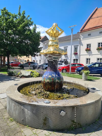 The image depicts a public fountain in Sankt Marein bei Neumarkt, Austria. The fountain features a golden statue of a figure with hands clasped overhead, atop a mosaic-covered spherical base. Water cascades from the statue into the basin, which is filled with water and small stones. The fountain is surrounded by a circular concrete structure with a perforated metal exterior. Paved walkways and greenery, including lavender bushes and a large tree, are visible around the fountain. In the background, buildings with tiled roofs and parked cars line a street. The sky is clear and blue with a few white clouds.