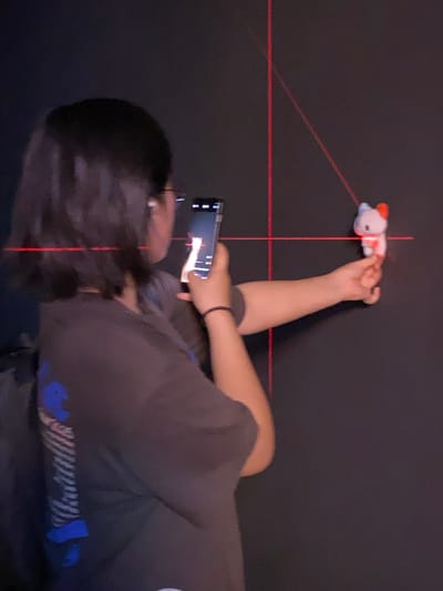 In the city of Jinniu District, China, an individual with dark hair, wearing a dark t-shirt and eyeglasses, is holding a smartphone in their right hand. The individual is looking at the phone, which displays a lit screen. In front of the individual, a small, stuffed toy with red and blue accents is being held in their left hand, positioned near a black wall. Two red laser lines are projected onto the wall, one vertical and one horizontal, intersecting each other. Another red laser line angles diagonally from the intersection point. The scene appears to be indoors, with the dark wall as the primary background.