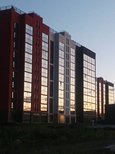 The image shows a row of modern, multi-story apartment buildings at dusk in Zapadny, Russia. The buildings are a mix of dark red, white, and black facades, all featuring large, reflective windows that mirror the sky and clouds. The windows suggest the apartments are newly constructed or under construction, as there are some visible gaps or open sections. Some windows have a dark, opaque appearance, while others reflect the warm hues of the sunset, creating a dynamic visual effect. The buildings have flat roofs with railings, and some appear to have balconies.

In the foreground, there is dark, unkempt grass and some debris, indicating the area might be an undeveloped lot or a construction site. The overall lighting suggests it is either sunrise or sunset, with a clear, pale blue sky dominating the upper portion of the image. The reflections in the windows are a prominent feature, capturing the dramatic colors of the sky, including oranges, yellows, and grays from the clouds. There are no visible people in the image, and the primary objects are the buildings and the natural landscape in the foreground. The scene evokes a sense of modern urban development against a natural backdrop, with the time of day lending a tranquil yet potentially transitional atmosphere. No visible text is present in the image.