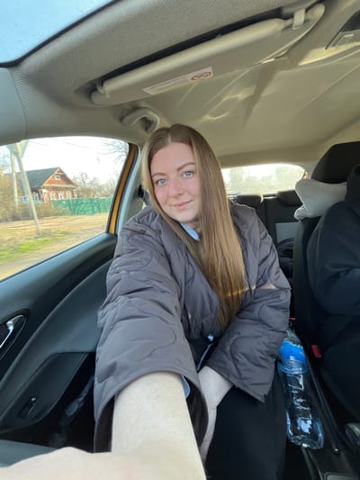 A young woman with long brown hair and blue eyes sits in the front passenger seat of a car. She is wearing a gray puffy jacket. A water bottle is visible in the back seat. Through the car window, a view of a street in Maksimkovo, Russia is visible. A red brick house with white trim stands alongside the road, suggesting a residential area. Grass and leafless trees are also visible. The car's interior is beige and black.