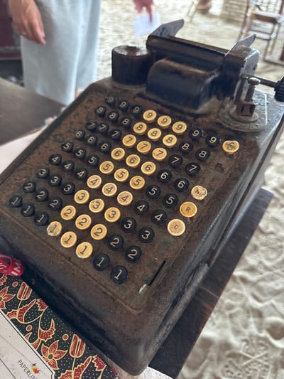 The image shows an antique adding machine, likely an "Underwood" brand, with a weathered, rusty surface and numerous keys. The keys are a mix of black and yellow, with some displaying numbers and others letters like 'N', 'R', and 'C'. A yellow key labeled "TOTAL" is visible on the upper right side of the keyboard. The machine is positioned on a dark wooden surface, which rests on a textured, light-colored fabric, possibly a tablecloth or rug, with a decorative patterned paper or fabric partially visible in the lower left corner. In the background, a person wearing a light blue garment is partially visible, suggesting the machine is in use or on display. The overall setting appears to be indoors, possibly in a shop or a home in the city of Gerokgak, Indonesia.