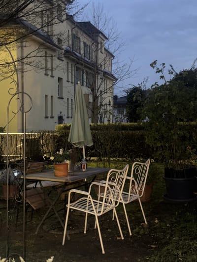 The image displays an outdoor garden area at dusk or early night in Bad Nauheim, Germany. In the immediate and mid-foreground, a rectangular wooden-topped folding table with a metal frame is situated on bare earth and sparse grass. A light green, closed patio umbrella is inserted through the table's center. Objects on the table include two stacked terracotta pots, a silver metal bucket containing white flowers, and a lidded German beer stein. Two white, ornate metal patio chairs with armrests are positioned to the right of the table, while a third wooden-slat folding chair is partially visible to the left. A dark metal garden arch with an attached, glowing solar light stands in the far left foreground.
In the background, a large, light-colored three-story building with a dark, multi-faceted sloped roof is visible. The building features multiple windows, many equipped with light-colored shutters. Several bare deciduous trees are positioned around and in front of the building. A dense, manicured hedge extends across the mid-background, creating a boundary. Beyond the hedge, additional distant structures are faintly visible under a dusky blue-grey sky. No individuals are present in the scene, and no actions or interactions are depicted.