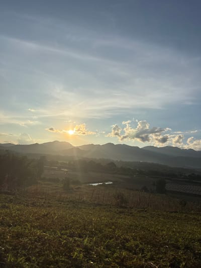 The image displays a panoramic landscape in Ban Mae Ping, Thailand, captured during either sunrise or sunset. The sun, a prominent golden orb, is low on the horizon, slightly left of center, partially obscured by bright, scattered cumulus clouds, and casting significant lens flares and visible light rays. A range of dark, undulating mountains stretches across the midground, silhouetted against the illuminated sky and partially veiled by atmospheric haze. Below the mountain range, a valley contains agricultural fields with varying shades of green and brown vegetation, interspersed with two visible small, dark bodies of water, likely ponds or paddies. Faint structural outlines are discernible in the mid-distance on the right. The foreground comprises a sloping field covered in green and dry brown grasses and low-lying plants, illuminated by the warm, directional light. No individuals or specific activities are observed.
