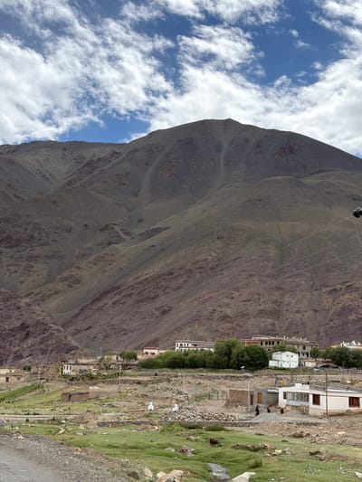 The image depicts a mountain village in Mahe, India. A large, barren, brown mountain dominates the background, with stark, eroded slopes marked by numerous gullies and ravines. The sky above is blue with scattered white clouds. In the mid-ground, a cluster of buildings forms the village. These structures vary in size and style, with some appearing older and made of mud or stone, while others are more modern with white and light-colored facades and visible windows. Green trees and vegetation surround the village. The foreground shows a grassy, uneven landscape dotted with rocks and small streams, with a gravel path leading into the scene from the lower left. Several people are visible in the village, some appearing to be engaged in construction or other daily activities. Power lines are also visible, stretching across the scene. The overall impression is of a remote, mountainous settlement.