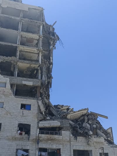 A partially destroyed, multi-story building stands against a clear blue sky. The building appears to be made of light-colored brick and concrete. Large sections of the upper floors have collapsed, revealing exposed structural beams, concrete debris, and twisted metal. Some of the lower floors are intact, showing windows, some with blue frames, and balconies with metal railings. Clothes hang from some of the windows, suggesting that people may still be inhabiting parts of the building. Rubble and debris are piled at the base of the structure.