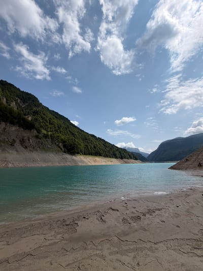 The image depicts a wide, tranquil body of turquoise water, likely a lake, surrounded by steep, forested mountainsides. The water's edge is marked by a dry, sandy shore with visible mud cracks and footprints, suggesting a receding water level. The sky above is a clear blue, dotted with scattered white clouds, casting soft shadows on the landscape. The mountains on the left are densely covered with dark green trees, their slopes appearing rugged and rocky in places. On the right, another mountain slope is visible, also covered in vegetation. In the distance, the landscape opens up to reveal a valley with more mountains under a hazy sky. The scene evokes a sense of natural beauty and peacefulness, typical of a mountain lake setting. The location is identified as Sauris / Zahre, Italy, a mountainous area known for its lakes and natural landscapes.