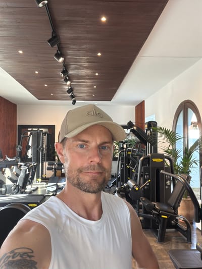 A man wearing a white sleeveless shirt and a beige baseball cap with the word "alo" embroidered on it is taking a selfie in a gym. He has blue eyes and a light beard. The gym has a modern design with a wooden ceiling and track lighting. Various weight machines, including Technogym equipment, are visible in the background, along with a treadmill and a large potted plant. The location is Sant Joan de Labritja, Spain.