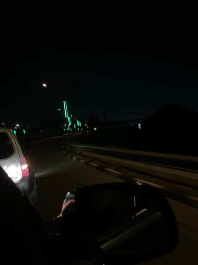 A nighttime image taken from inside a vehicle. The vehicle's side mirror is visible in the right part of the frame, reflecting the dark environment. A vehicle's rear lights appear at the lower center. In the left background, there are illuminated green bars on a tower, appearing to be telecommunications. The moon is barely visible in the left side of the image.
Given the location is Jiru, Nigeria, the tower could be a cell phone tower or other broadcast tower.