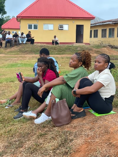 The image shows a group of people sitting on the ground outside a yellow building with a red roof. Several people are in a "piggyback" like position. The location is Ise - Ekiti, Nigeria. The environment appears to be a grassy area or field. Several people are sitting on the ground near the buildings. The group of people in the foreground are sitting very close together.