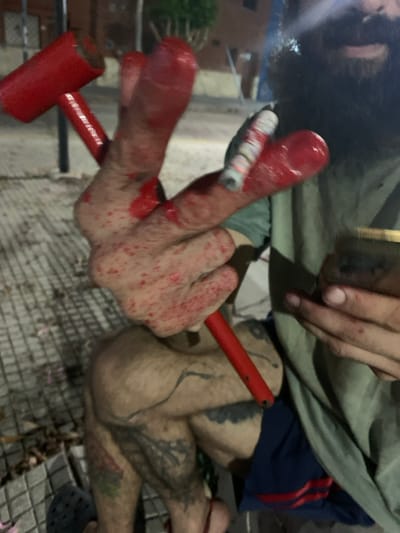 An individual is depicted in a close-up, low-light setting on patterned grey pavement in Villa Bernardo de Monteagudo, Argentina. The subject's right hand (viewer's left) is prominently displayed, heavily coated in a wet, red substance consistent with blood, particularly on the extended index and middle fingers. Smaller red splatter marks are also visible across the dorsal surface of this hand. This hand grasps the red handle of a red-painted hammer, identified as a mallet or sledgehammer. A lit cigarette, actively smoking, is held between the middle and ring fingers of the same hand. The subject's left hand (viewer's right) holds a small, brass-colored cylindrical object and also exhibits some red substance. The lower portion of the subject's face is obscured by a dark, full beard. Below the waist, blue shorts with red stripes are visible, and both legs are tattooed, with dark sandals worn on the feet. The immediate ground features dried leaves and debris. The background reveals an urban environment at night, characterized by indistinct buildings and artificial light sources.