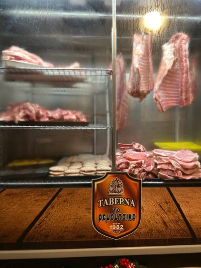 The image displays a refrigerated glass display case filled with various fresh meat cuts, likely inside a butcher shop or a taverna in Agios Dimitrios, Greece. The perspective is slightly angled, looking into the case.

On the top shelf of the display case, several large portions of meat, appearing to be racks of ribs (possibly pork or lamb), are neatly arranged. Below them, on the middle shelf, more similar rib cuts are visible, along with a stack of what looks like pork chops or steaks on the right side. The bottom shelf holds more meat, including rows of what appear to be sausages or ground meat patties, some individually wrapped in plastic. The glass of the display case has some condensation or streaks, and a bright light source is reflected in the upper left corner.

To the right of the display case, a decorative wooden sign is prominently featured. The text on the sign is in Greek and reads "TABEPNA" (TAVERNA), "TO PEMOYTΣIKO" (TO REMOUTSIKO), and "από το 1982" (since 1982), indicating the name and establishment year of the eatery. The background behind the sign features warm-toned wooden paneling. A small, festive decoration, possibly a part of a Christmas wreath with red berries, is faintly visible on the far right edge of the frame, suggesting the time of year might be around the holiday season. The overall scene suggests a traditional Greek restaurant or butcher showcasing its fresh meat offerings.
