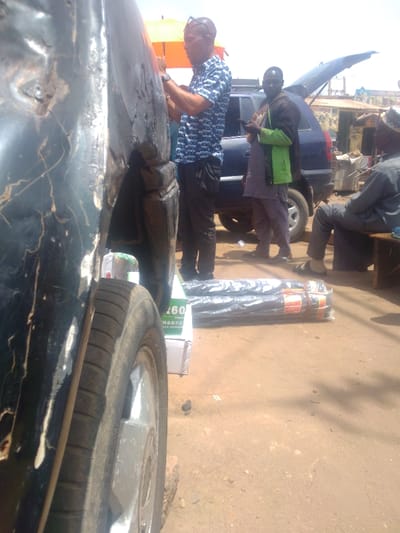 The image presents an outdoor scene in Mista Ali, Nigeria, characterized by an unpaved, reddish-brown dirt ground and bright daylight.
In the immediate foreground, the front left fender and wheel of a dark-colored vehicle are prominently visible. The fender exhibits significant damage, including multiple dents and extensive paint chipping that exposes a darker underlayer. The tire appears worn.
Midground elements include three individuals and two vehicles. A man, wearing a blue and white patterned short-sleeved shirt, dark trousers, and sunglasses on his head, stands facing and interacting with the damaged dark vehicle's side. To his right, a second man stands, holding a smartphone, dressed in a dark long-sleeved top, a bright green vest, and light-colored trousers. Behind these two individuals, a dark blue SUV is parked with its rear hatch open. An orange umbrella is visible above the head of the man in the patterned shirt.
On the ground in the midground, two bundles of dark material, rolled and encased in clear plastic with red and yellow securing tape, are positioned alongside a white box with partially visible green lettering. Further to the right, an older man in dark attire and a cap sits on a wooden bench, facing left.
The background reveals a series of rudimentary commercial structures with corrugated metal roofs, indicating a market or roadside vendor area. The sky above is bright and clear.