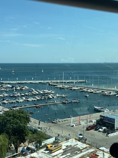 This media file captures an aerial view of a marina in Gdynia, Poland. The scene is dominated by numerous sailboats and yachts docked along a pier, with a vast expanse of calm, blue water stretching into the distance. The sky is clear and bright, suggesting a sunny day.

Several people are visible on the pier, some walking and others gathered near boats, implying activity related to boating or a maritime event. A few small boats are also in the water, adding to the dynamic feel of the marina. On the land side, a building with a dark roof and windows is present, and some construction activity is evident with scaffolding and building materials. A yellow vehicle, possibly a utility cart or small truck, is parked near the building.

The image appears to be taken during daylight hours. The presence of numerous sailboats and the general atmosphere suggest a recreational or possibly a competitive sailing event. The location cues of "Gdynia, Poland" are provided, which, combined with the visual of a bustling marina, paints a picture of a vibrant coastal city. No specific emotions are readily discernible from the static image, but the bright conditions and lively marina suggest a pleasant and active day. No legible text is clearly identifiable within the image.