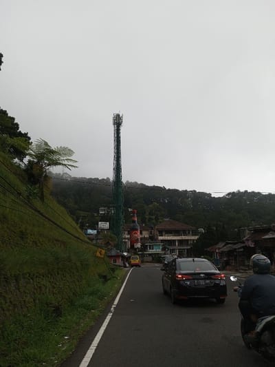 The scene depicts an uphill view along an asphalt road under an overcast sky, purportedly in the city of Phoxay, Laos. In the immediate foreground, the paved road features a solid white line on its right edge. A black four-door sedan, bearing Indonesian license plate "B 1522 SNU," is centered in the right lane, moving away from the viewer. To the right of the sedan, the rear of a person on a scooter, wearing a dark jacket and light helmet, is partially visible, also heading forward.

To the left of the road, a steep, grass and dirt-covered embankment ascends, marked by multiple dark cables running diagonally upward. A large fern-like plant is situated on this slope. In the mid-ground, a tall, green lattice telecommunications tower stands prominently to the left of the road. Below the tower and slightly to its right, a large billboard shaped like an "ABC Kecap Manis" bottle is visible, positioned near other structures. A yellow mini-van is parked near the base of these signs. Several multi-story buildings, including one with a reddish-brown tiled roof and light-colored, weathered walls, are nestled into the hillside behind the tower and billboard. More modest buildings line the right side of the road further into the mid-ground. The background consists of densely forested hills receding into the hazy, light grey sky.