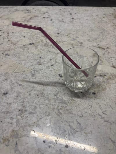 A burgundy drinking straw with a flexible joint rests diagonally across a clear, faceted glass tumbler. The straw's end is angled upwards and to the left, while the other end disappears into the tumbler. The glass sits on a speckled, light-colored countertop, likely granite, with dark inclusions and lighter veining. A faint shadow of the straw is cast to the left onto the countertop. The background is dark and out of focus. The scene is indoors and appears to be a domestic setting in Ghukasavan, Armenia.