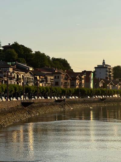 The image depicts a waterfront scene in Zumaia, Spain, during what appears to be the golden hour. In the foreground, a body of water, likely a river or bay, shows ripples and reflections of sunlight. A stone embankment lines the water's edge, leading to a walkway or street. Behind the embankment, a row of buildings with traditional architecture, featuring red-tiled roofs and balconies, is visible. These buildings are interspersed with lush green trees. Further in the background, a taller, modern building with a lighthouse-like structure on top stands out. The sky is a soft gradient of pale blue and yellow, indicating either sunrise or sunset. The scene is quiet and appears to be devoid of any visible people or immediate activity, suggesting a peaceful moment in the city.