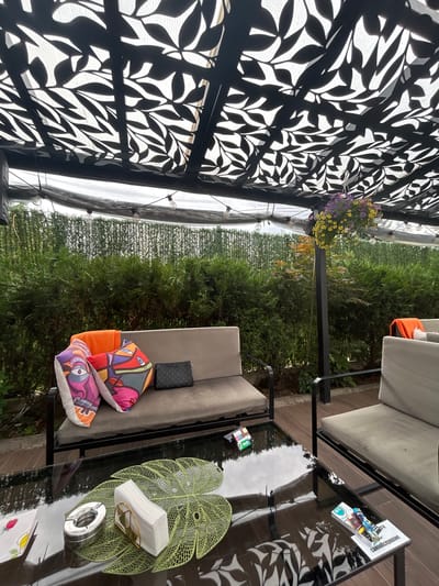 This image depicts an outdoor seating area, likely in Dolna banya, Bulgaria. The scene is set under a black perforated canopy that casts a leaf-like shadow pattern. In the foreground, to the left, is a section of this canopy. To the right, a black glass-topped table sits on a patio. On the table, there are several items including a metal ashtray, a decorative green leaf-shaped placemat holding a stack of white napkins with a golden clip, and a magazine or brochure. Reflecting in the table's surface are trees and the sky, suggesting an outdoor setting.
Behind the table, a cushioned outdoor sofa is visible, with a dark patterned clutch bag placed on its backrest. Two brightly colored decorative pillows with abstract designs, one featuring an eye-like motif, rest on the sofa. Further to the right, another section of a sofa is visible, with an orange cushion placed on it.
Beyond the seating area, a dense green hedge forms a backdrop. A hanging basket filled with yellow and purple flowers is suspended from a black metal pole that supports the canopy. String lights with exposed bulbs are also visible, partially draped along the edge of the canopy. The overall impression is a relaxed outdoor space, likely used for lounging, with a focus on decorative elements and comfort. The lighting suggests it could be daytime, with diffused light filtering through the canopy. There are no people visible in the image.