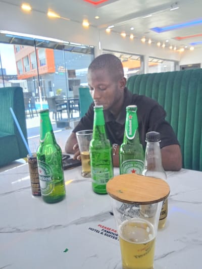 The image shows a man sitting at a table in a restaurant, holding a glass of beer in his hand. There are three unopened bottles of beer on the table, one of which is a Heineken, and a glass of what appears to be a soft drink on the table. The man is wearing a black T-shirt and his head is bowed. The table is made of white marble. The restaurant is bright and modern, with large windows and a green booth behind the man. The location is in Ujevwu, Nigeria, at the coordinates 5.472 latitude, 5.818 longitude. There is a sign on the table that reads "Please patronize Famous Hotel & Suites." The man is seated alone, looking at something on the table.