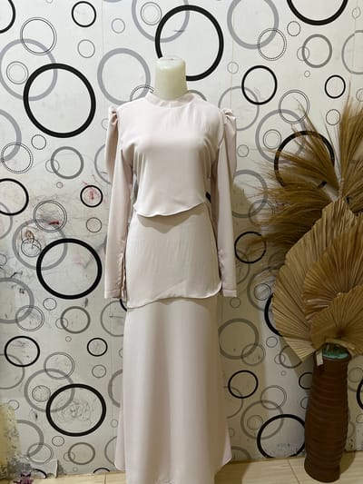 A light pink, two-piece outfit, likely a blouse and skirt, is displayed on a mannequin against a patterned wallpaper featuring black and grey circles. To the right of the mannequin, a brown vase holds dried palm fronds, adding a touch of natural decor to the scene. The background wallpaper is visually striking, with a mix of solid and dotted circles in varying sizes, creating a dynamic and modern aesthetic. The outfit itself appears to be made of a smooth, possibly satin-like material, with the blouse featuring long sleeves and a layered detail. The skirt is long and flows down, completing the elegant silhouette. The overall setting suggests a retail or display space, possibly in Payacut, Indonesia, given the context of the request.