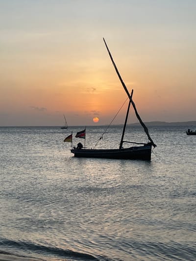 A long, narrow boat sits on calm water, possibly a dhow given its tall, angled mast. Two small flags hang from the stern. The sun sets or rises in the background, casting a yellow-orange glow on the horizon and sky. The water reflects the sky's colors, with slight ripples on the surface. The scene exudes tranquility and a sense of remote location.