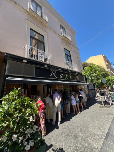 The image shows a street scene in Sorrento, Italy, on a sunny day. A building with a pale pink facade dominates the foreground, with a gelato shop called "Raki" visible at street level. The shop has a black awning and large glass windows displaying the interior. A line of people, dressed in casual summer clothing, is waiting outside the shop. Some individuals are looking at the menu or the gelato inside, while others are standing and chatting. A large potted plant with green leaves and white flowers is in the lower left corner of the frame, partially obscuring the gelato shop. The street pavement is made of dark grey stone tiles. In the background, another building, painted yellow, is visible, along with trees and a clear blue sky. A street lamp and some street signs can also be seen. The overall atmosphere is one of a busy tourist destination on a pleasant day.