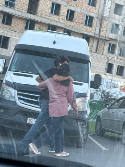 The media file shows two individuals, a man and a woman, embracing each other in the street. The man is wearing a black t-shirt, and the woman is wearing a pinkish-purple shirt and blue jeans. They appear to be standing in front of a white van, with their faces obscured. The embrace suggests a moment of affection or comfort.
The scene is set on a street with a large, multi-story building under construction in the background, characterized by exposed brick and incomplete window frames. To the right of the van, another vehicle, possibly a grey SUV, is visible. The ground is asphalt, with a white arrow painted on it, indicating a directional marking on the road.
The lighting suggests it might be daytime, perhaps late afternoon or early evening, given the relatively soft light and shadows. There are no strong indicators of specific weather conditions.
A small white license plate with red text "05 KG" is visible on the back of the van, providing a location cue. The overall atmosphere seems to be one of intimacy amidst a busy, urban environment, possibly indicating a personal moment shared between the individuals. The presence of the construction site suggests the location is an area undergoing development.