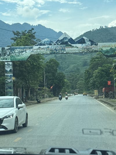 The image depicts a road in Minh Tân Commune, Vietnam, as seen from within a vehicle. A sign for the "Dong Van Karst Plateau UNESCO Global Geopark" is visible. Several motorbikes are traveling along the road, along with at least one car. Trees and mountains form the backdrop. A Vietnamese flag is also visible.