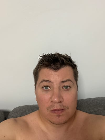 A man is photographed from the chest up, against a plain white wall. He has short, dark, messy hair and light green eyes. His skin is fair with some redness on his cheeks. He has a thin mustache and a small goatee. He is shirtless and appears to be sitting on a grey sofa. The scene is indoors, with no indication of location within the city of Benimàmet-beniferri, Spain.