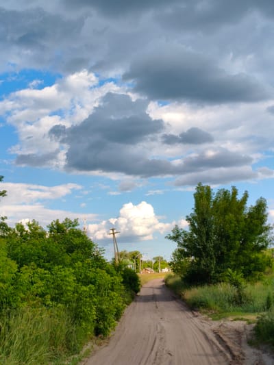 A dirt road curves through a rural landscape under a dramatic sky filled with dark, heavy clouds interspersed with patches of bright blue. On either side of the road, lush green foliage and tall grass create a sense of abundance and natural growth. In the distance, a transmission tower and power lines are visible, hinting at the proximity of civilization. The presence of a lone wooden utility pole with insulators further accentuates the rural setting. The overall impression is one of a serene yet potentially volatile day, with the changing weather adding a dynamic element to the scene. The image appears to be taken in daylight, likely late afternoon given the cloud formations and lighting. The location is identified as Matiashivka, Ukraine. There are no people or animals visible in the image, and no text is present.