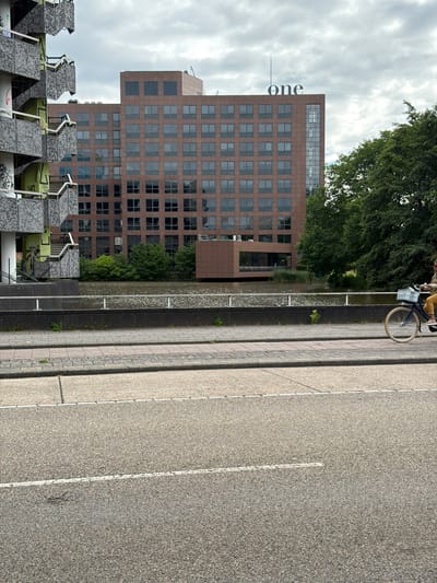 A person rides a bicycle with a light blue basket on the right side of the image, on a paved walkway. The walkway is adjacent to a body of water. In the background, a large, multi-story brown building with many windows stands. The building has the word "one" in large white letters on its upper facade. To the left of the brown building is a gray, multi-level structure with external staircases, appearing somewhat dilapidated and covered in graffiti. Trees line the edge of the water between the walkway and the buildings. The sky is overcast with clouds. The foreground shows a section of asphalt road with a white line. This scene is located along the Amstel River in Amstelveen, Netherlands, near the office building commonly known as "The One" or "De Kantoorkolos" for its distinctive appearance and the prominent signage.