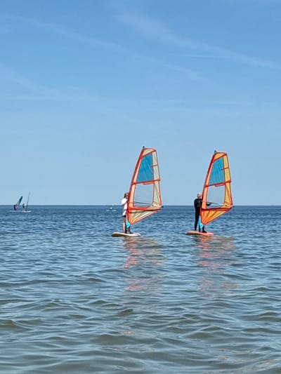 The image depicts a scene of windsurfing on a body of water, likely a lake or the sea, under a clear blue sky with faint wispy clouds. In the foreground, two windsurfers are prominently featured. The windsurfer on the right is wearing a black wetsuit and is holding onto the boom of their sail, which is a vibrant orange and yellow with blue accents. They are standing on a windsurf board. To the left, another windsurfer, wearing a white t-shirt and dark shorts, is also on a windsurf board with a similarly colored sail. In the background, several other windsurfers are visible further out on the water, appearing as smaller figures with their sails. The water has a rippled surface, reflecting the sky and the sails. The horizon is a clear line separating the water from the sky. The scene suggests a recreational activity taking place on a sunny day. There is no indication of the city of Rewa, Poland in this image.