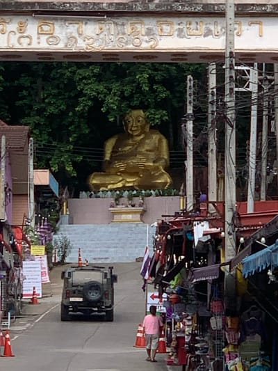 This is a medium-shot, slightly elevated, outdoor, full-color photograph taken in the Mae Sai District of Thailand. The scene depicts a road in a market area, with a large golden Buddha statue visible in the background. The statue is seated and positioned on a raised platform with stairs leading up to it. The statue is framed by lush green trees and an ornate archway inscribed with what appear to be religious or decorative motifs.
In the foreground, a dark-colored SUV is driving down the road. The vehicle has a spare tire mounted on the back. On either side of the road, there are market stalls with awnings and various goods displayed. Several orange traffic cones are placed along the road.
To the left of the SUV, a person wearing a pink shirt and light-colored shorts is standing on the sidewalk, facing away from the camera. There are also Thai flags visible, attached to some of the market stalls. The overall impression is a busy roadside market scene with a prominent religious landmark in the distance.