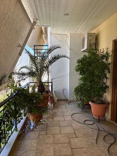 The image depicts a balcony in Ilion, Greece, with a tiled floor and a white wooden ceiling. Two large potted plants, a palm tree and a ficus, are prominent. An air conditioning unit is mounted on the wall. A retractable awning extends from the ceiling, partially covering the balcony. A garden hose is coiled on the floor, and a spigot is visible on the wall. A metal railing lines the edge of the balcony, overlooking a street with buildings and a clear blue sky. The overall scene suggests a residential balcony setting, possibly for leisure or gardening.